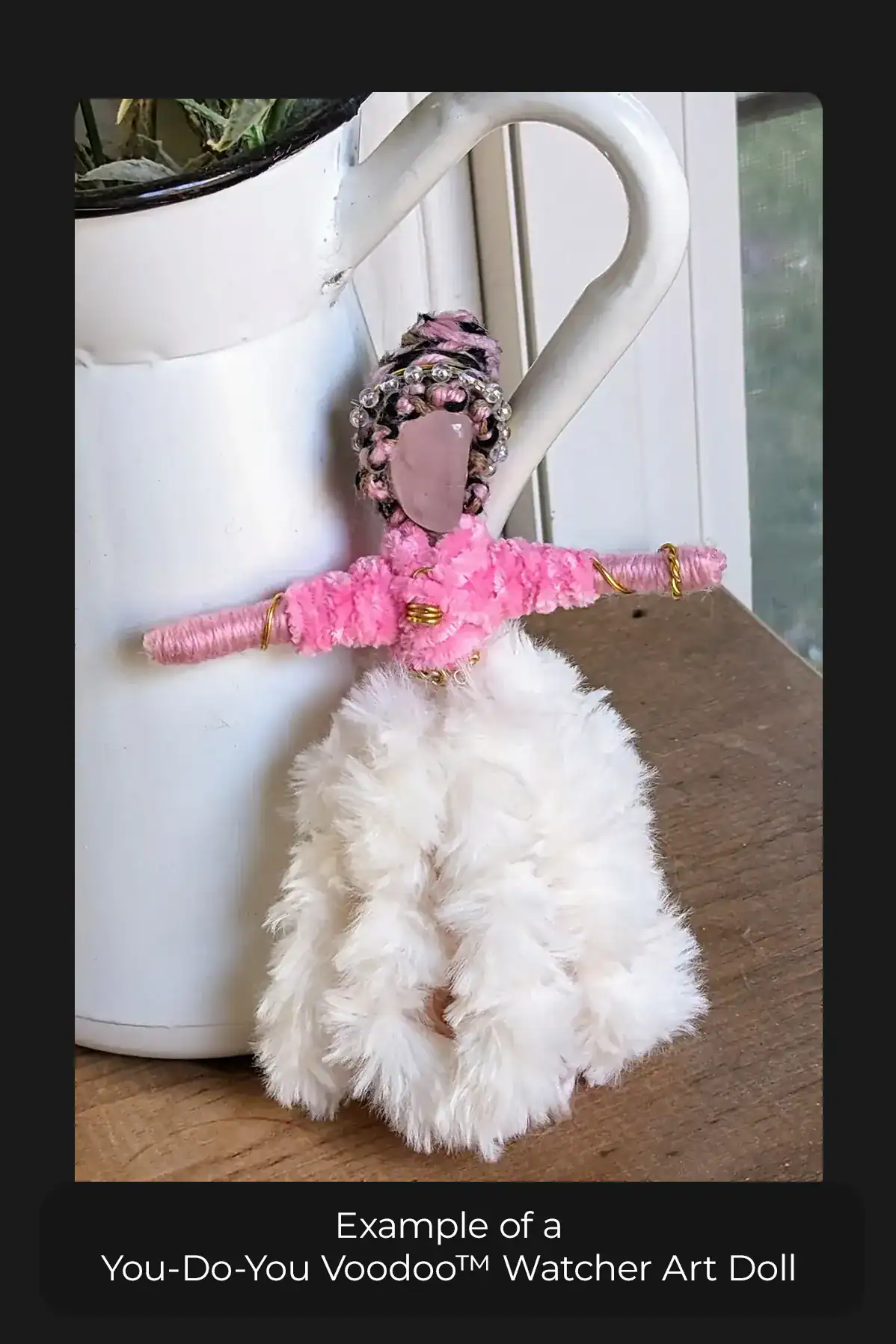 A You-Do-You Voodoo™ Watcher art doll dressed in a pink top, furry skirt, and jewelry. Her black, pink and brown hair is intricately braided. She's standing by a decorative pitcher.
