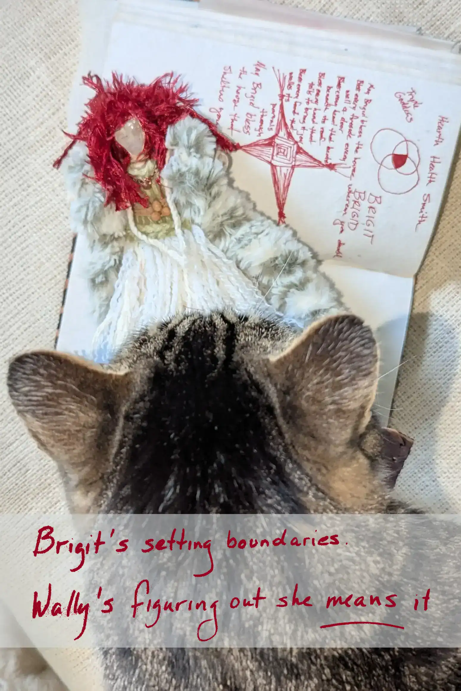 Tabby cat inspecting Brigit You-Do-You Voodoo™ doll resting on an open journal with a Brigid’s cross, red yarn hair, faux fur, and white yarn skirt visible.