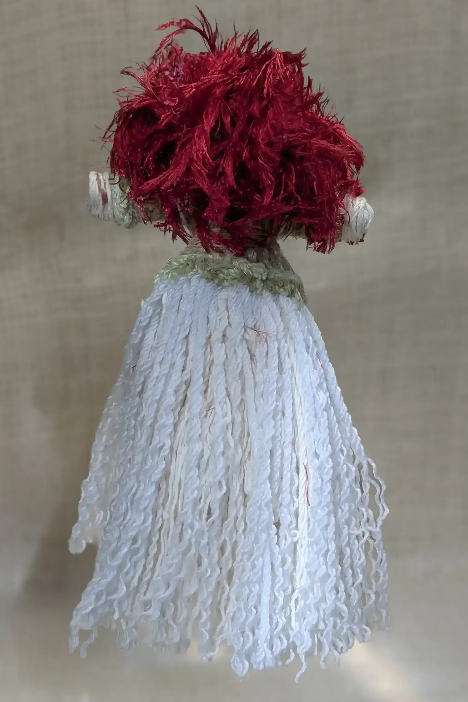Back view of handmade Brigit You-Do-You Voodoo™ doll with red yarn hair, soft green top, and full white yarn skirt, standing against a neutral background.