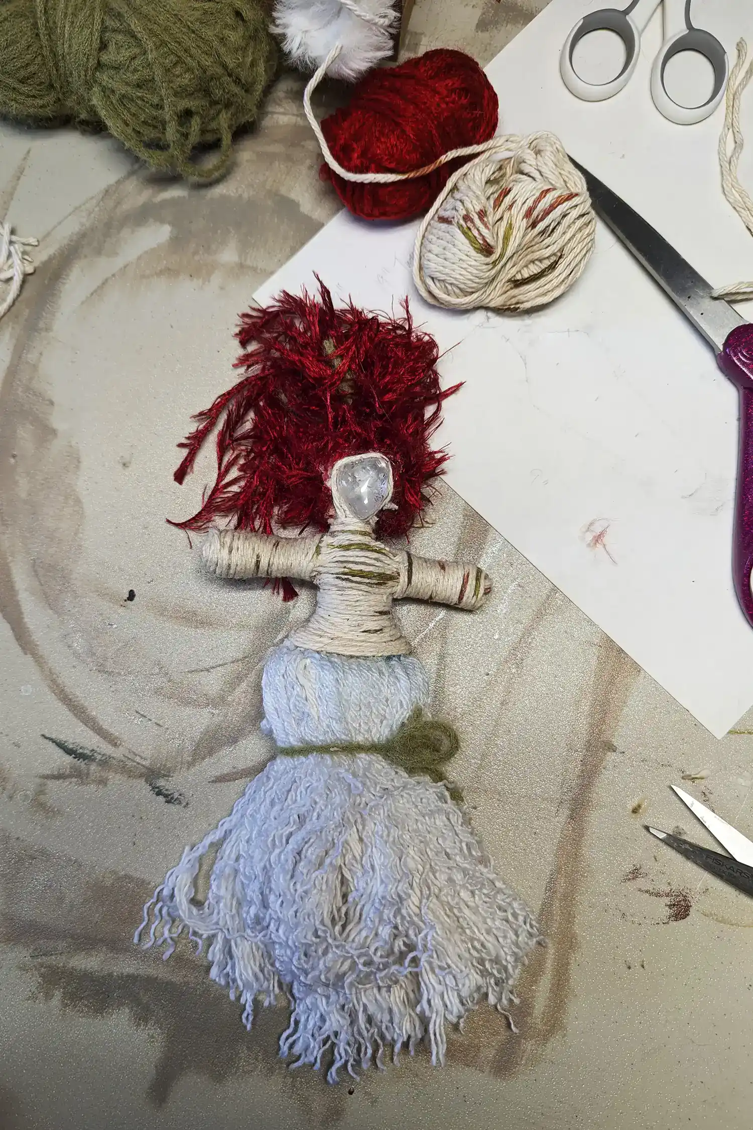 Partially completed Brigit You-Do-You Voodoo™ doll with clear quartz head and red yarn hair, lying on artist workspace with yarn, scissors, and fiber remnants.