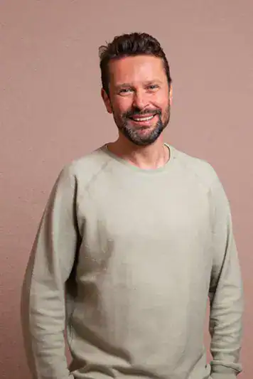 Smiling man with a beard and casual beige sweater posing against a neutral background.
