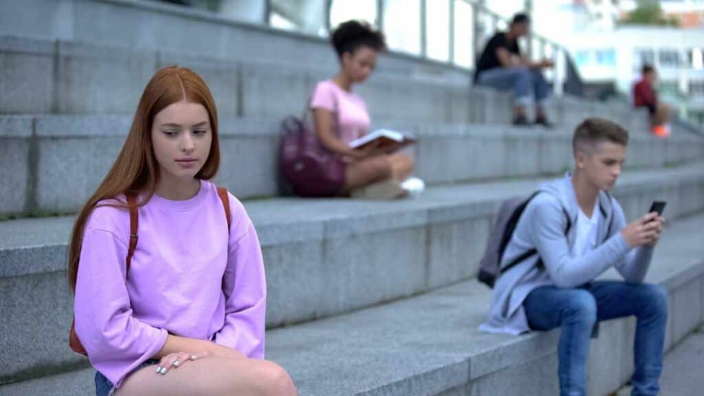 Sad teen girl sitting alone on school steps, while peers are nearby, illustrating social withdrawal, a sign of adolescent depression.