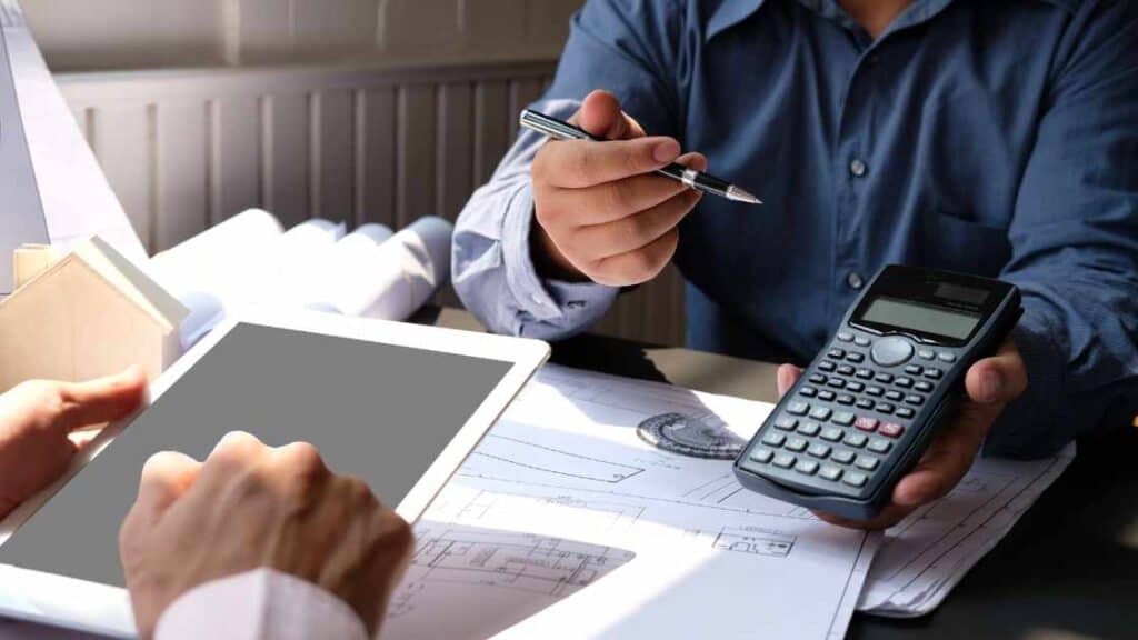 Two people discussing teen depression treatment costs using a calculator, tablet, and architectural cost documents.
