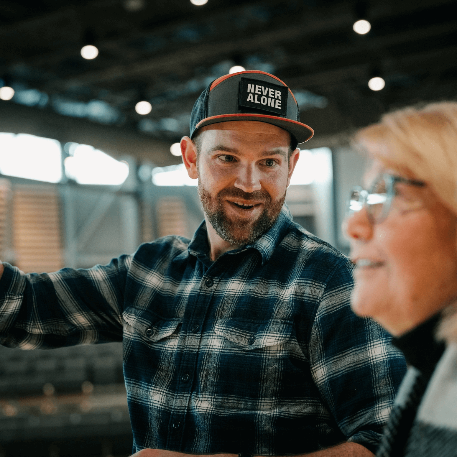 A smiling man wearing a "Never Alone" cap engaging in a friendly conversation with an older woman, both appearing happy and connected.