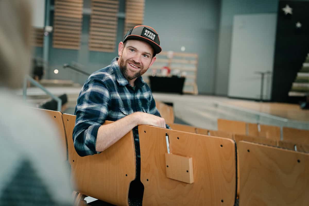 A smiling young man with a beard, and ballcap seated in an auditorium.