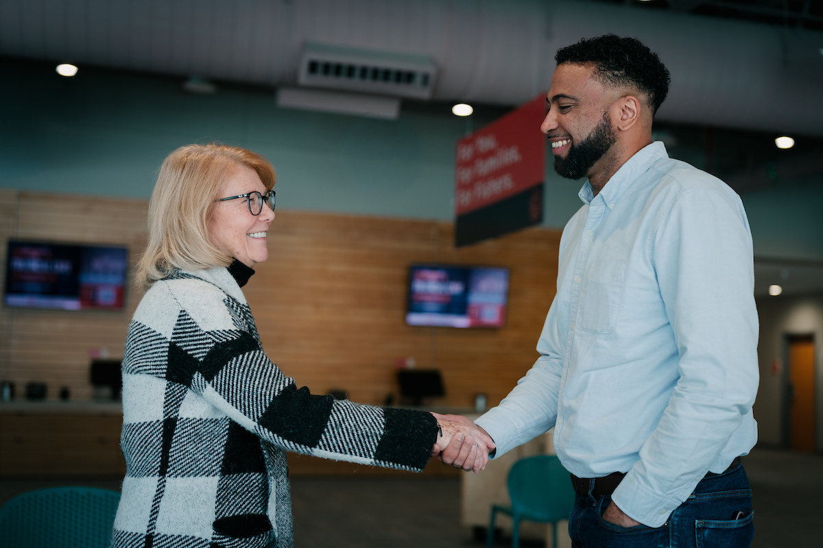A woman and a man shaking hands in a modern office environment, smiling and making eye contact, with digital screens and a wooden wall in the background.