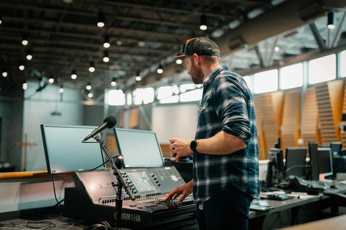 An image of a man wearing a plaid shirt and cap working with sound mixing equipment in a spacious, well-lit auditorium