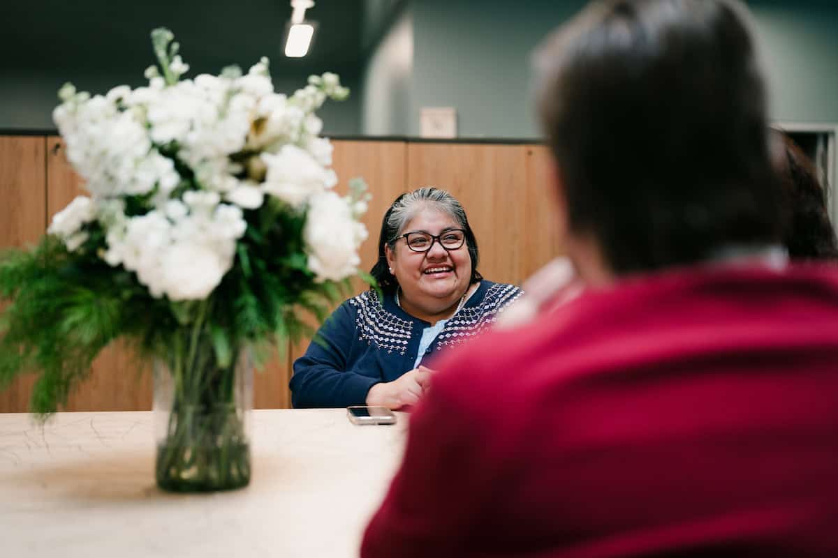 A woman with glasses and a blue sweater smiling and talking to another person in a red top