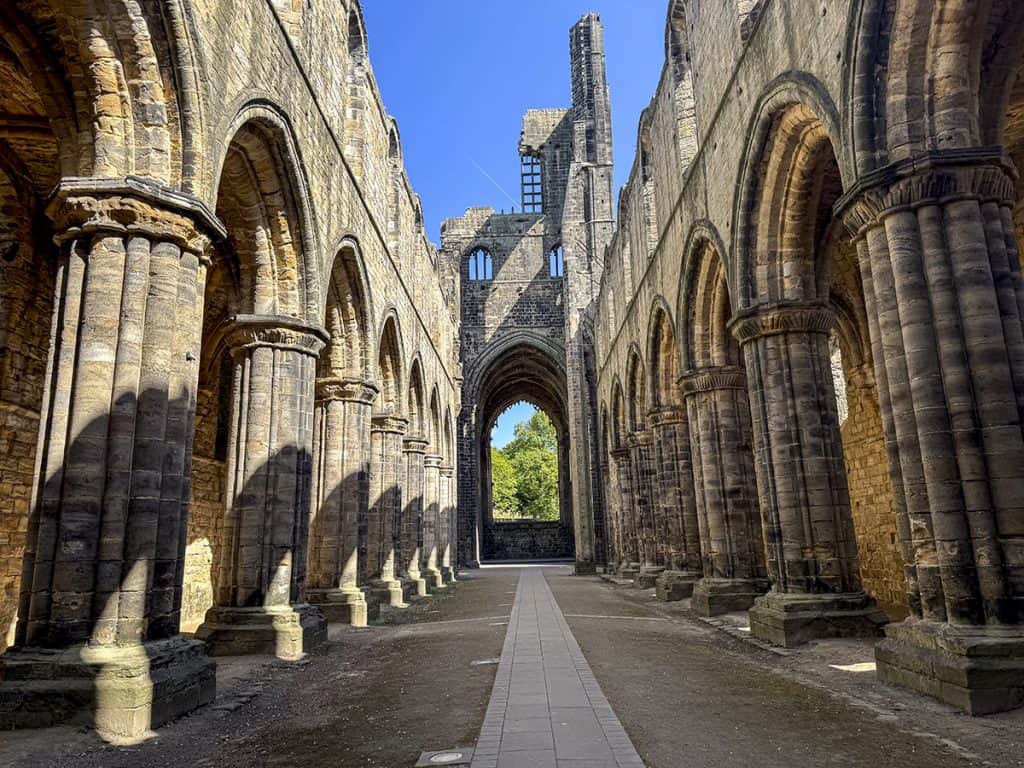 Interior of Kirkstall Abbey Church, with rows of stone arcade columns lining a central paved path through the nave, an uneven mix of earth, stones, and gravel either side, and a large arched window visible at the far end.