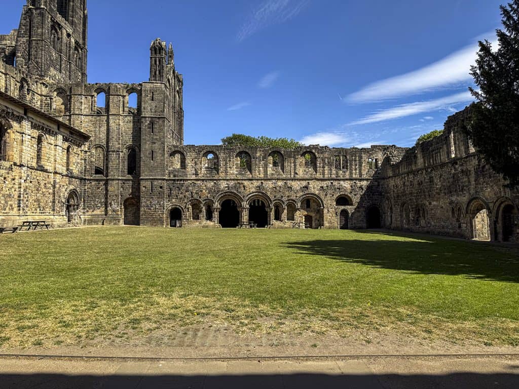 Exterior of Kirkstall Abbey's Cloister, with a grass courtyard surrounded by a tarmac path and ruined stone walls.