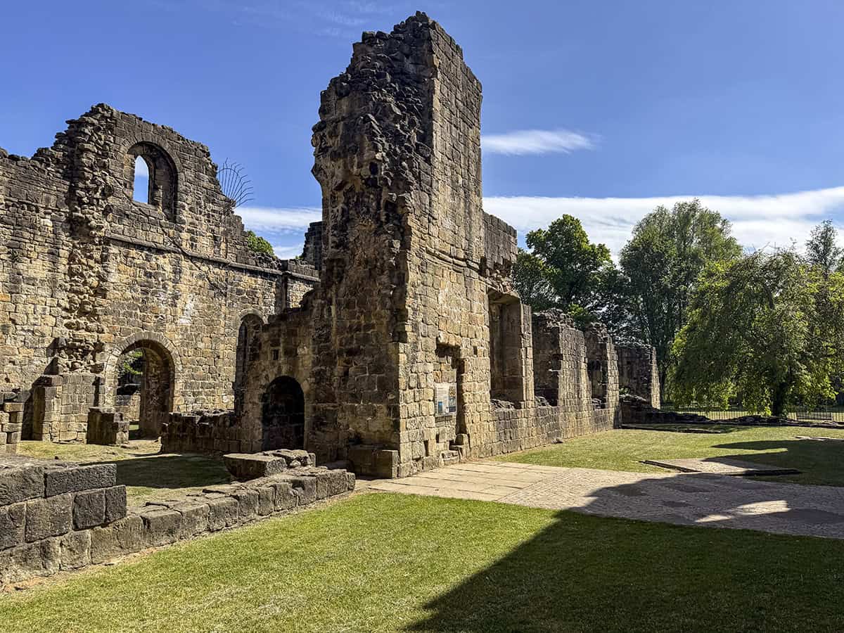 Exterior of Kirkstall Abbey Refectory, with ruined stone walls and a mix of grass, stone paving, and surviving patterned monastic floor tiles underfoot.