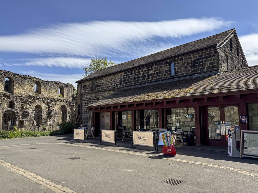 Exterior of Kirkstall Abbey's visitor centre, a glass-fronted stone building, with a tarmac pathway and large automatic double doors.
