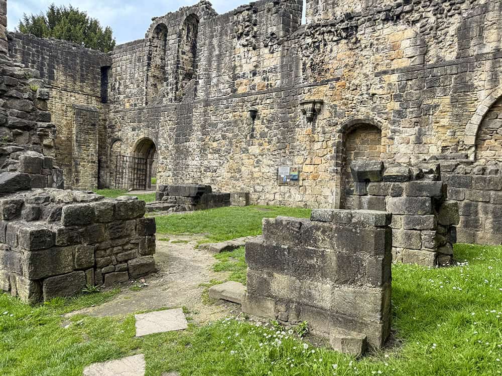 Exterior of Kirkstall Abbey Refectory ruins, with a mixed pathway of stone, grass, and gravel, and the Cloister entrance visible beyond.