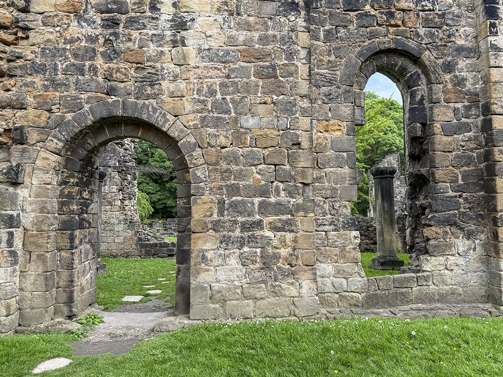 Exterior of Kirkstall Abbey's ruined stone wall between the Refectory and Infirmary, with a stepping-stone path leading through a doorway arch.