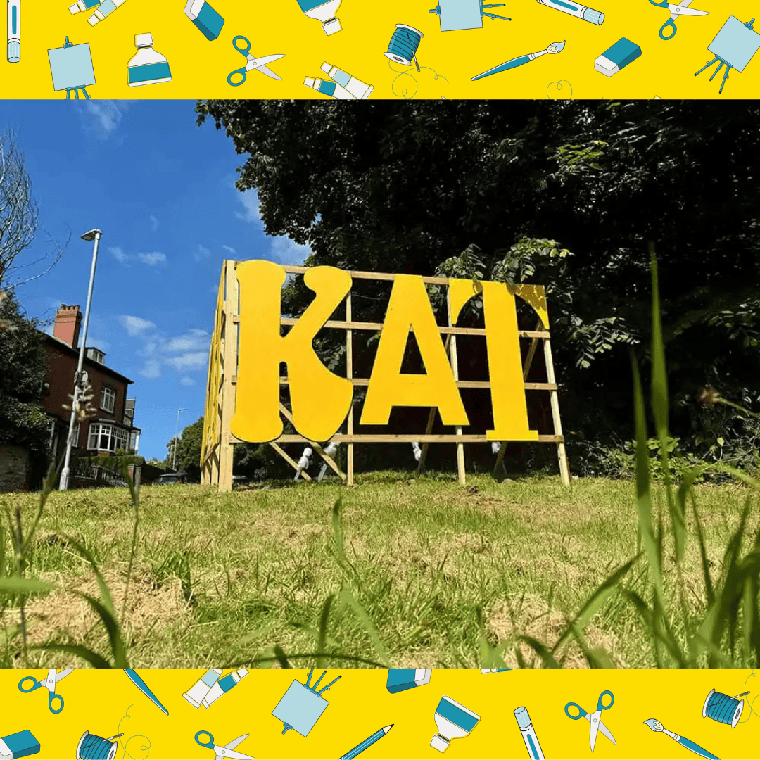 The KAT sign. Large bright yellow letters, built out of wood. They stand on a grass mound .