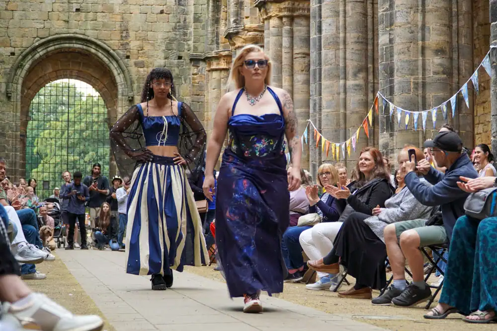 Two models walk together: one in a sheer black-sleeved top with a striped blue-and-cream skirt, the other in a strapless navy dress with a floral bodice and starry skirt.