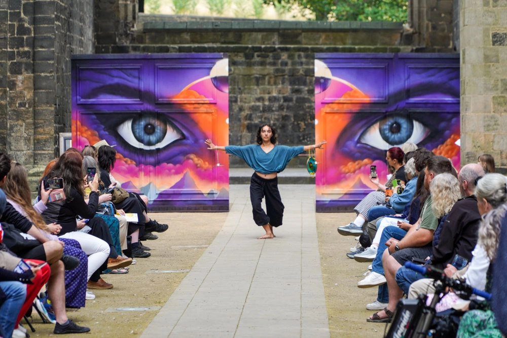 A performer in a teal wrap top and black wide-leg trousers dances barefoot on the catwalk, arms outstretched, in front of a large painted eye mural.
