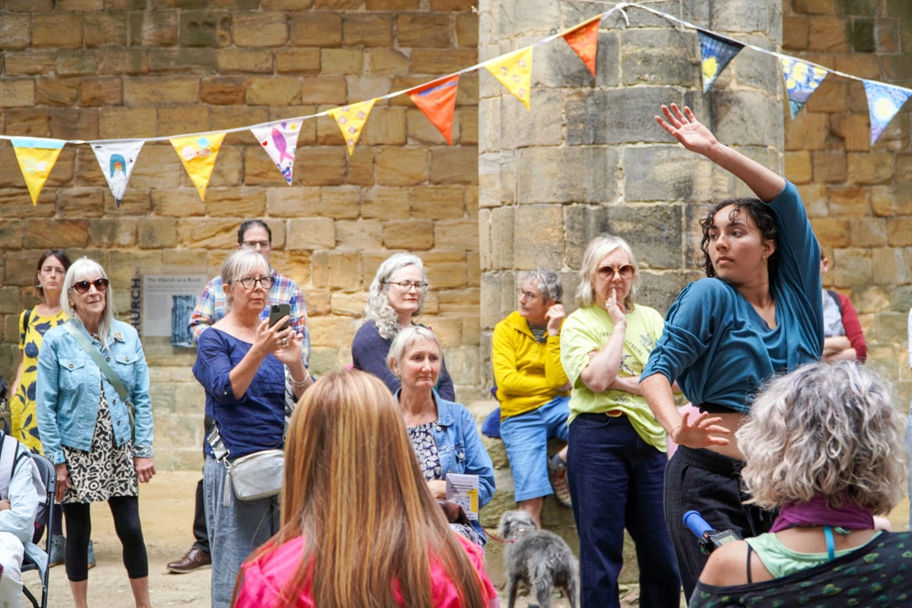 A barefoot performer in a teal top and black trousers is surrounded by an audience.