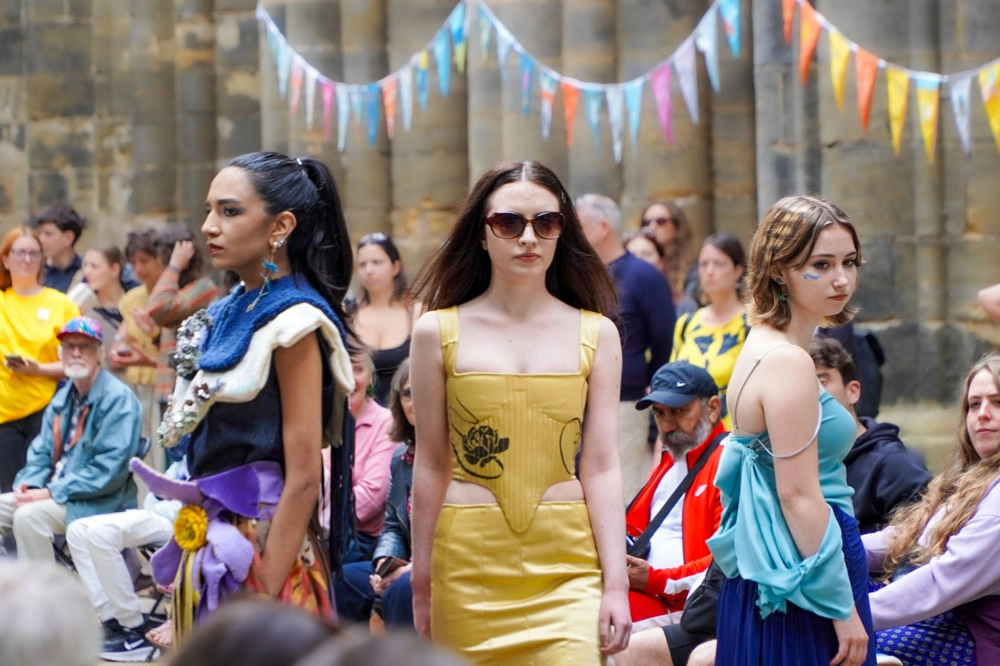 Three models on the catwalk: one in a navy outfit with a sequinned shoulder piece and purple flower detail, one in a yellow corset dress with black floral print, and one in teal draped fabric over a navy dress.