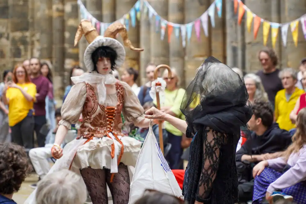 Two costumed models: one in a rust-and-cream corseted dress with a ram-horn headpiece, the other veiled in black lace, holding a white paper lantern.