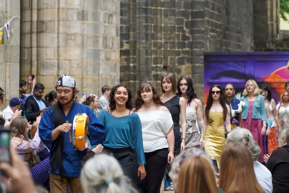 A group of smiling models walk the catwalk together, led by a drummer in a blue tunic playing a frame drum.