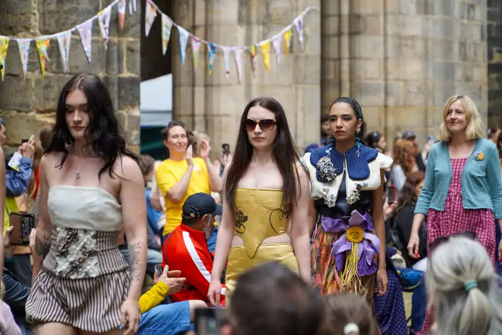 Three models on the catwalk: one in a striped fabric top and skirt, one in a yellow corset dress, and one in a white felted shoulder piece over a patterned skirt with a purple flower.