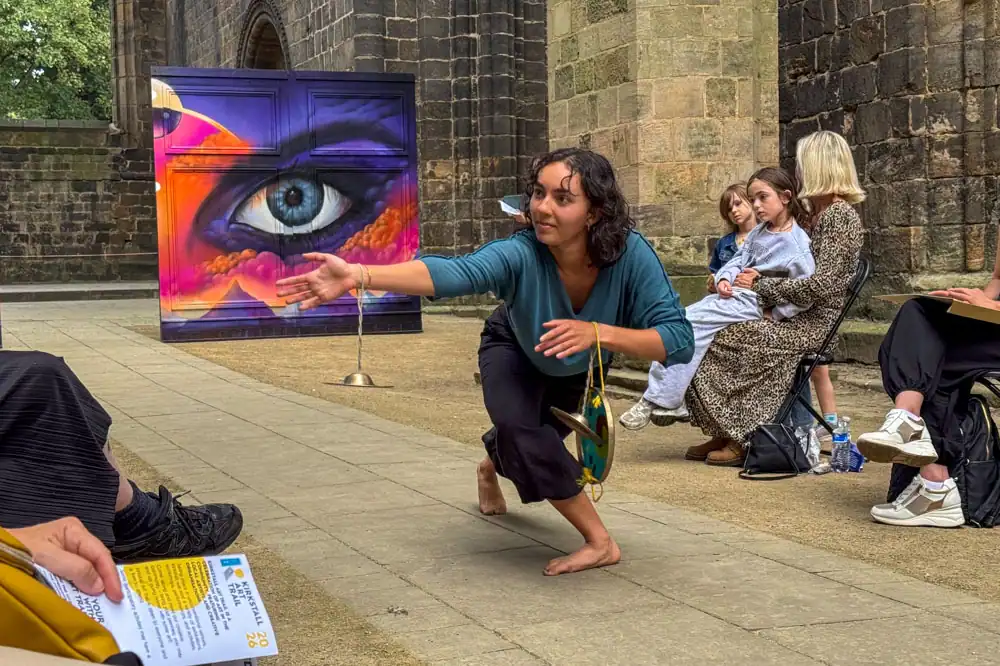 A barefoot performer in a teal top and black trousers crouches and reaches out, small cymbals hanging from her wrist, in front of a painted eye mural.