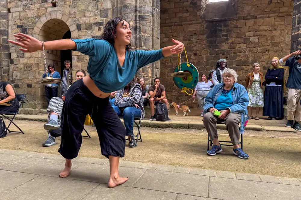 A barefoot performer in a teal top and black trousers, her body contorted with arms outstretched, holding a small cymbal.