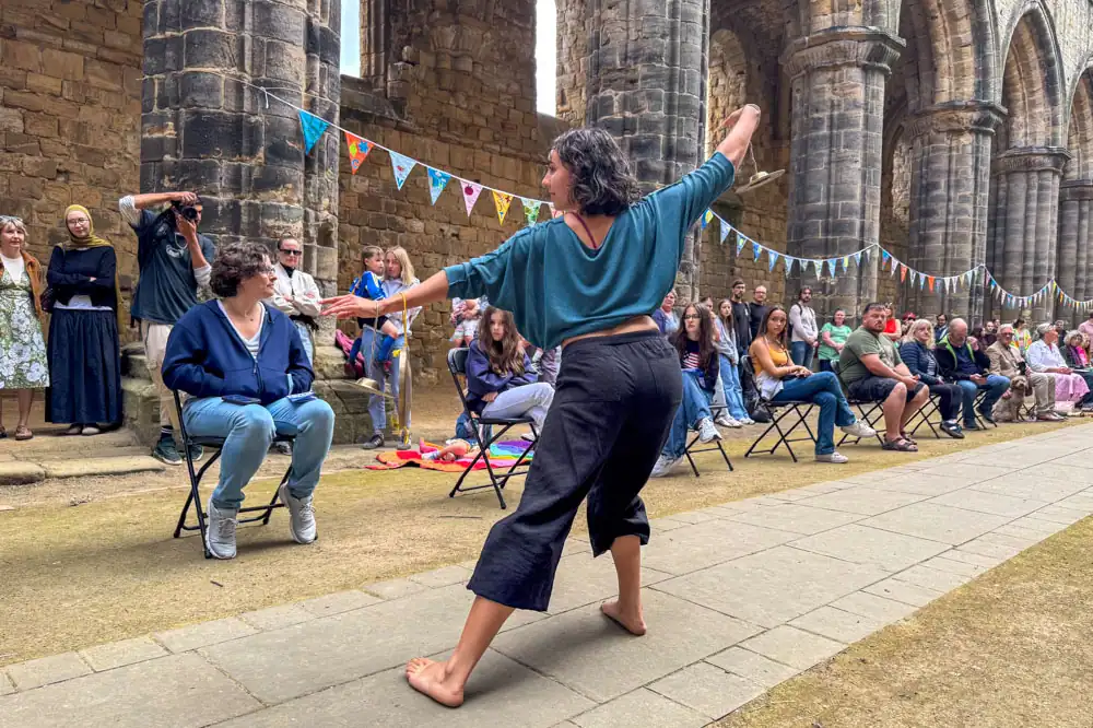 A barefoot performer in a teal top and black trousers, her arm outstretched with a cymbal in hand.