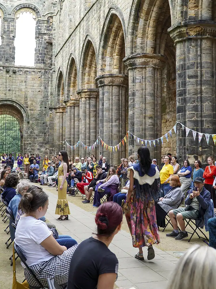 The fashion show catwalk showing the length of the nave, with bunting strung between stone columns and a packed audience on either side as models walk between them.