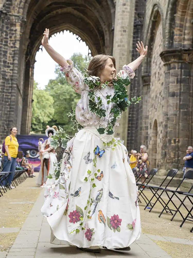 A model in a white floral blouse and long skirt, draped in ivy vines with butterfly and bird appliqués, poses with arms raised.