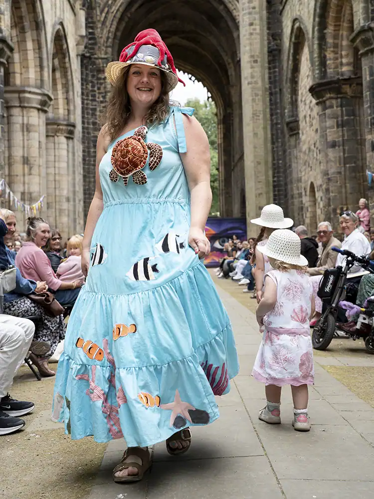 A model walks in a pale blue dress appliquéd with a turtle, fish, and starfish, wearing a straw hat topped with a red toy octopus.
