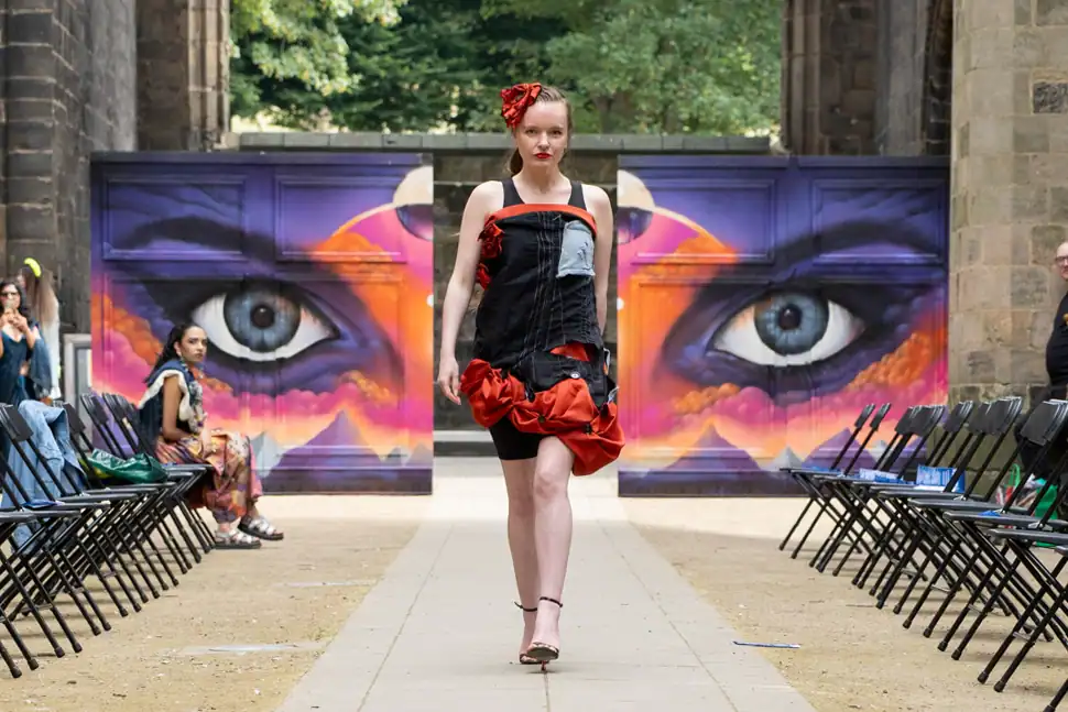A model in a black structured dress with red ruffle detailing and a red flower hair accessory walks the catwalk in front of a painted eye mural.