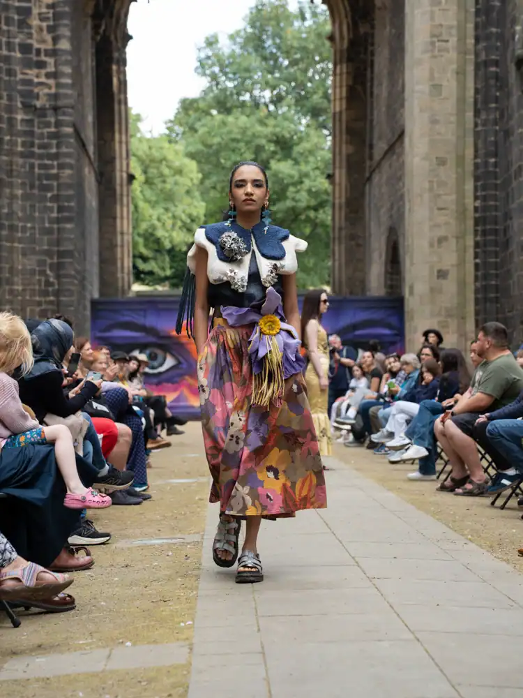 A model walks in a white felted cape-style shoulder piece over a floral patterned midi skirt with a tasselled purple flower.