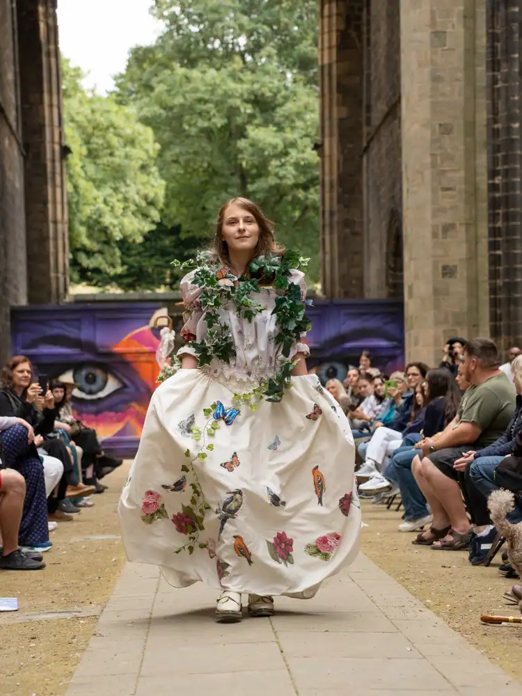 A model in a white floral blouse and long skirt, draped in ivy vines with butterfly and bird appliqués, walks the catwalk.
