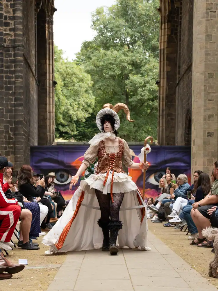 A costumed model walks the catwalk in a rust-and-cream corseted gown with a full cream skirt trimmed in orange, wearing a horned bonnet headpiece and holding a crook.
