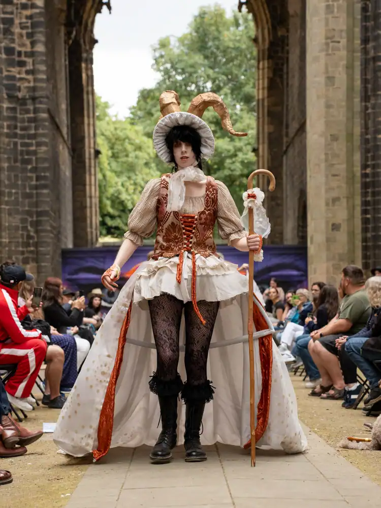 A costumed model stands in a rust-and-cream corseted gown with a full cream skirt, black lace tights, and boots, wearing a horned bonnet headpiece and holding a crook.