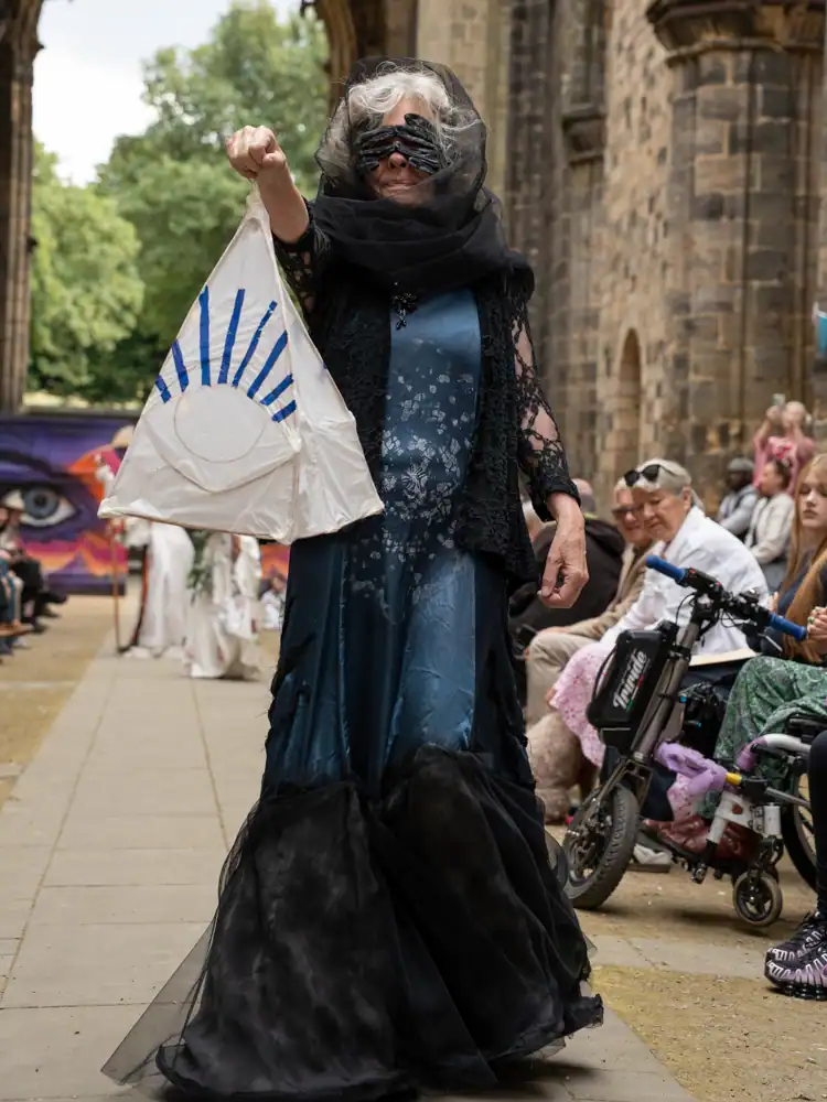 A costumed model in a black lace veil and shawl over a blue-grey gown with a black ruffled hem, holding a triangular paper lantern printed with an eye motif.
