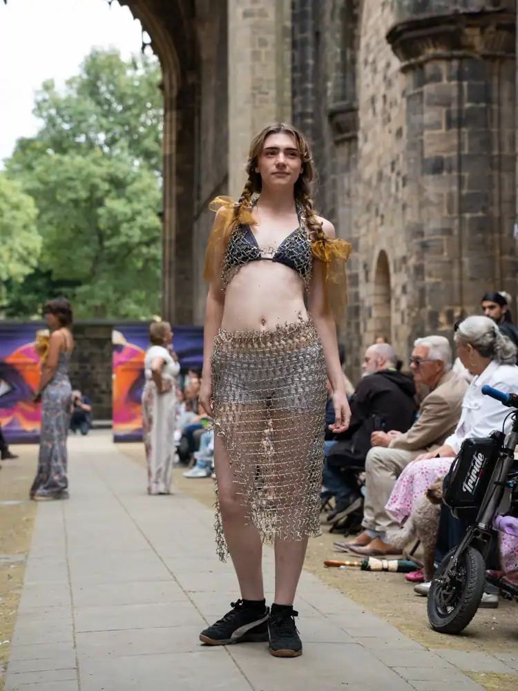 A model stands in a chainmail-style bikini top and matching draped skirt with dangling metal charms, with sheer orange bows in braided hair.