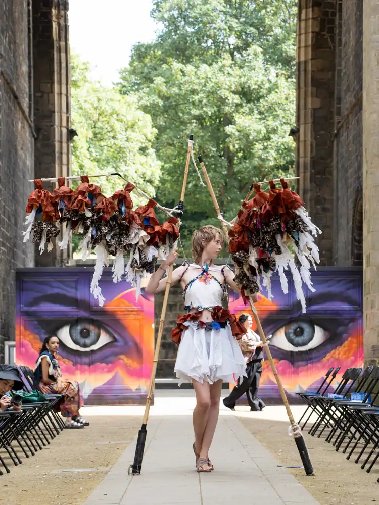 A model walks the catwalk in a white halter top and skirt with rust-coloured fabric trim, carrying two tall poles hung with rust and cream fabric strips resembling wings, barefoot.