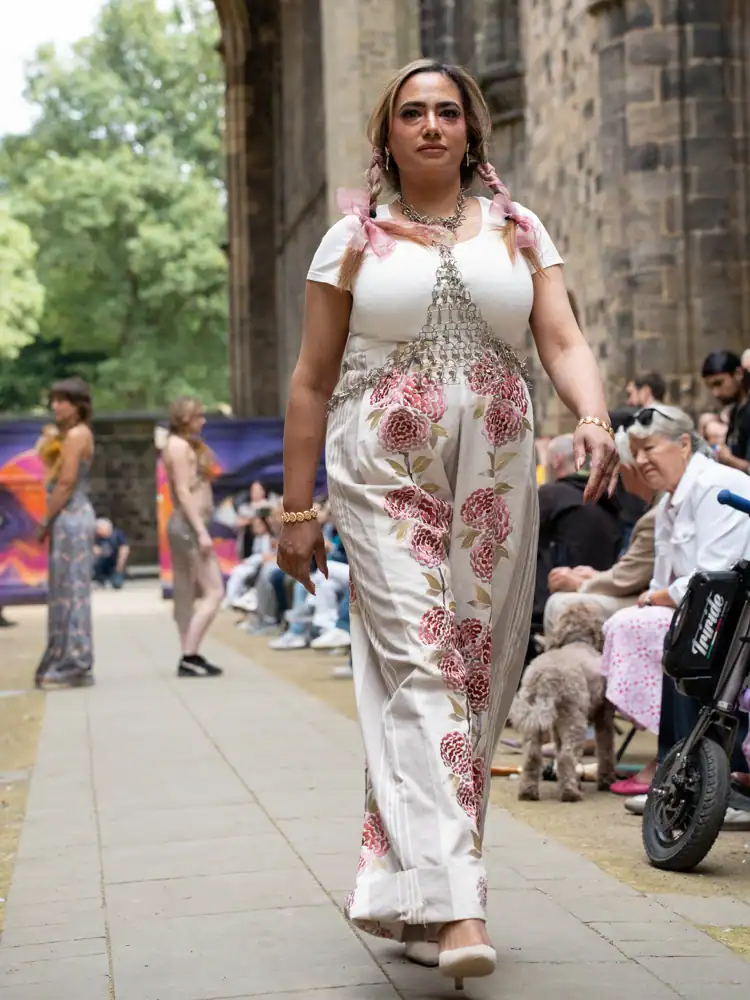 A model walks the catwalk in a white top and wide-leg trousers embroidered with large pink floral motifs, layered with a draped metal chain necklace.