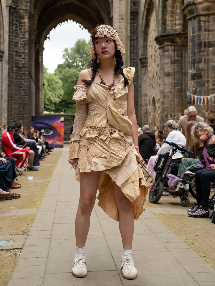 A model in a distressed, layered cream fabric dress with an asymmetric hem, a lace headscarf, and one crocheted sleeve.