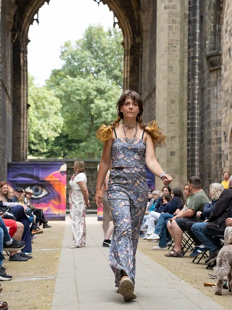 A model walks the catwalk in a blue floral-print cami top and matching wide-leg trousers, with sheer gold bows in braided hair.