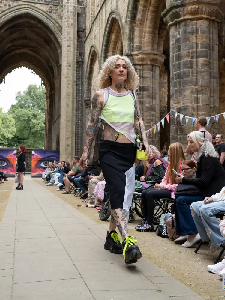 A model walks the catwalk in a neon green cropped top, sheer overlay skirt with white and black panels, and chunky black platform trainers with neon laces.