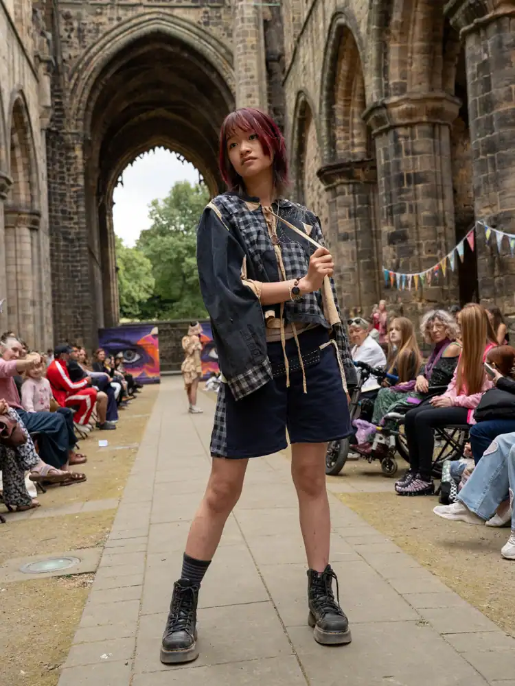 A model stands on the catwalk in an oversized checked shirt-jacket layered over a cream top, with navy shorts and chunky black boots.