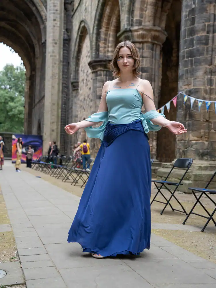 A model walks the catwalk in a pale blue-green off-shoulder top with draped sleeves and a navy floor-length skirt.