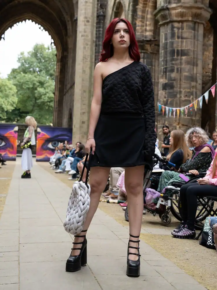 A model walks the catwalk in a black quilted one-shoulder top and matching mini skirt, carrying a chunky knit white bag, with strappy platform heels.