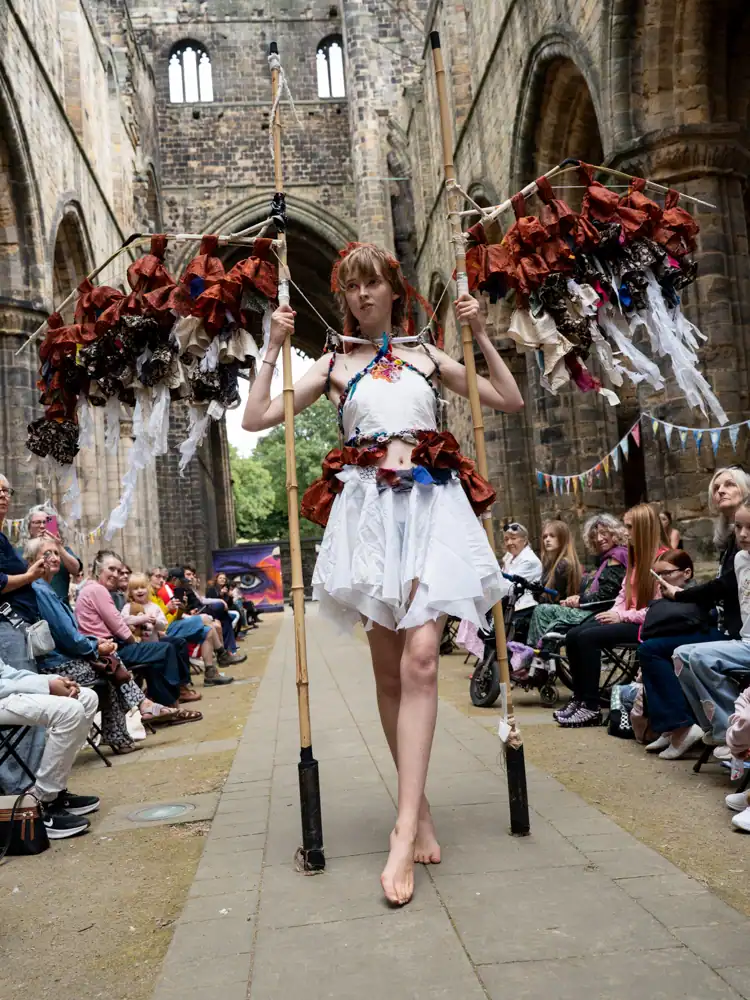 A model walks the catwalk in a white halter top and skirt with rust-coloured fabric trim, carrying two tall poles hung with rust and cream fabric strips resembling wings, barefoot.