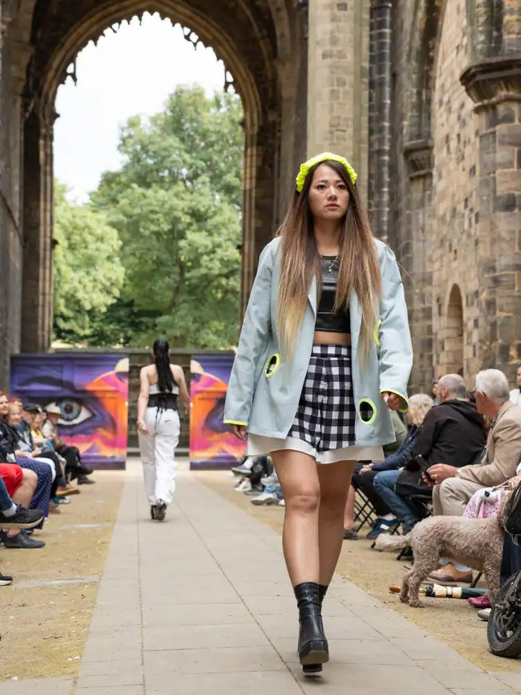 A model walks the catwalk in a pale blue jacket with neon yellow circular cutouts, a black crop top, and a black-and-white gingham skirt over a white underskirt, wearing a neon flower headband.