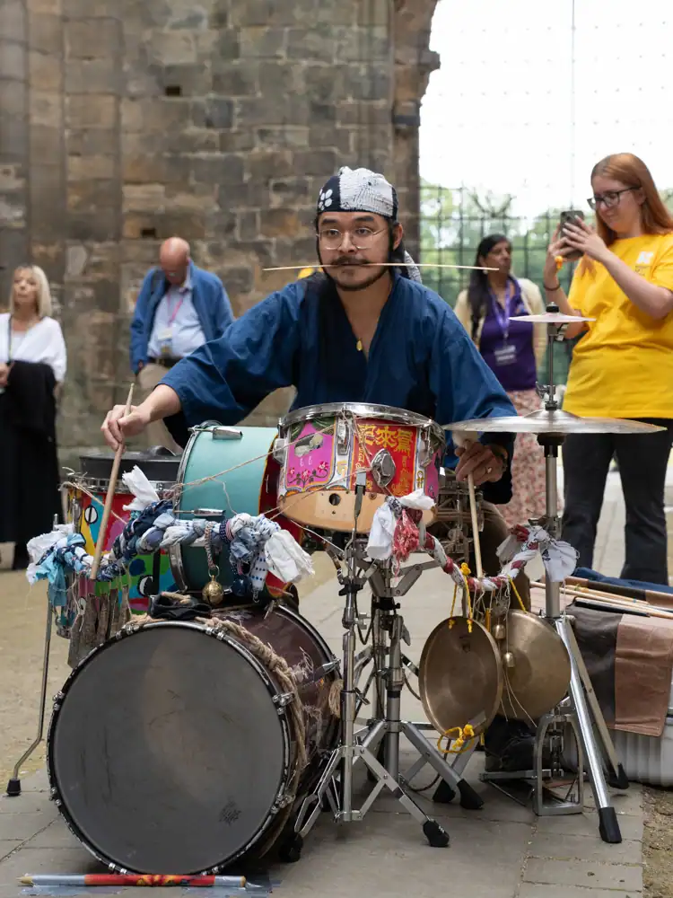 A musician in a blue robe and headscarf plays a decorated drum kit with cymbals, a drumstick held between his teeth.